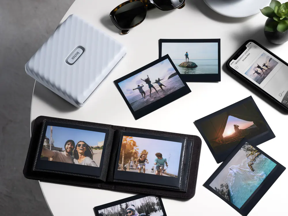 Фотопринтер Fujifilm Instax Link Wide на столе рядом с напечатанными широкоформатными снимками, альбомом и смартфоном с приложением instax