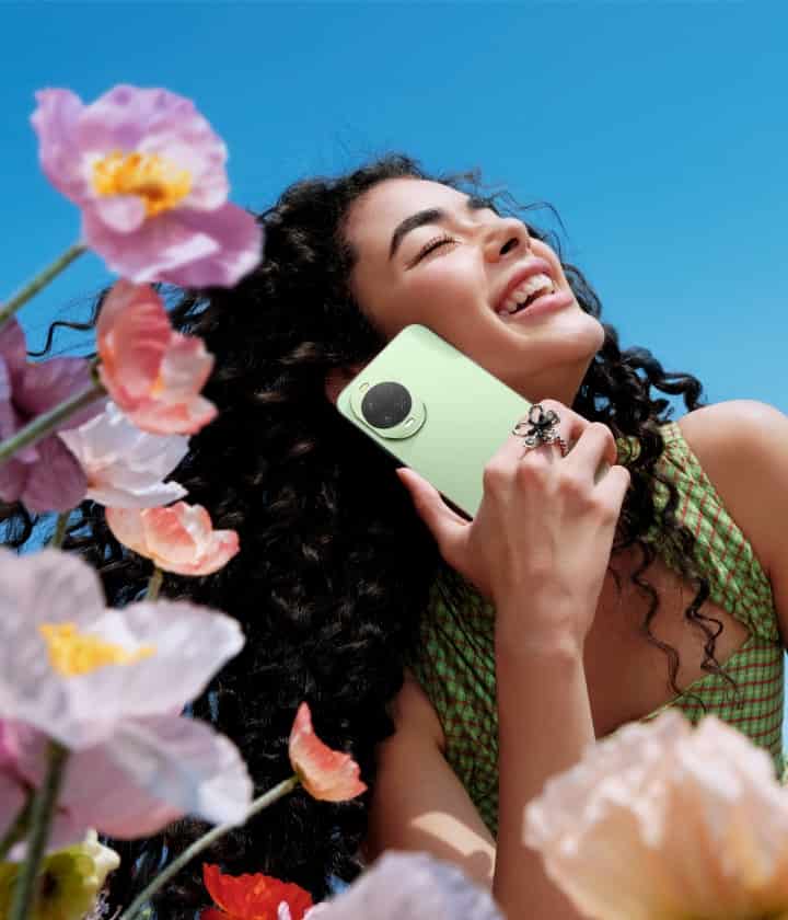 Девушка держит у щеки зелёный Huawei Nova 15 на фоне цветов